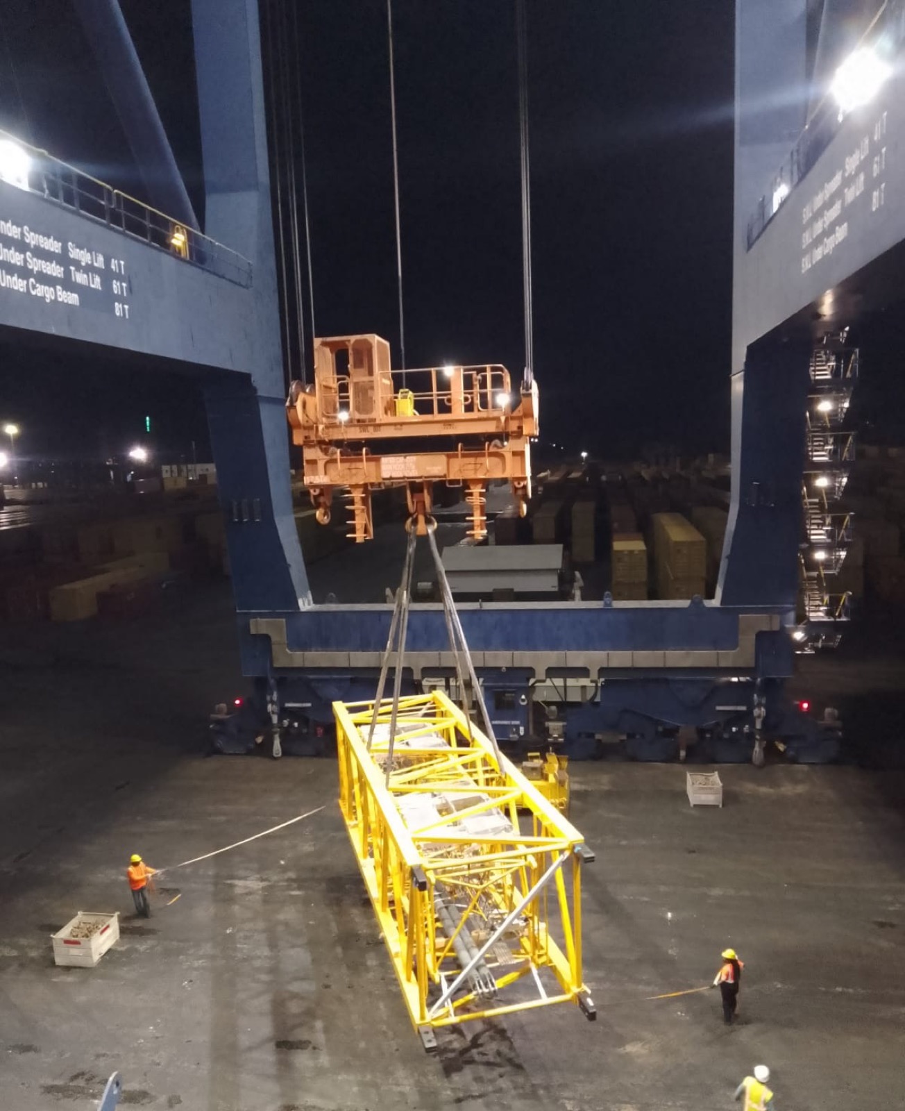 Crane lowering a yellow spreader beam into a ship's cargo hold at night