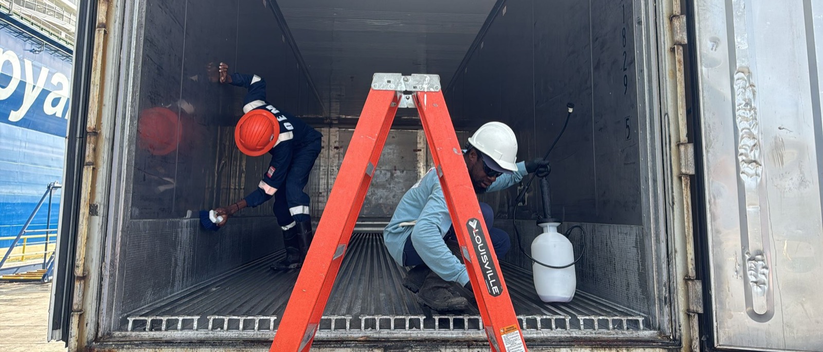 Two technicians cleaning a container interior with brushes and sprayer